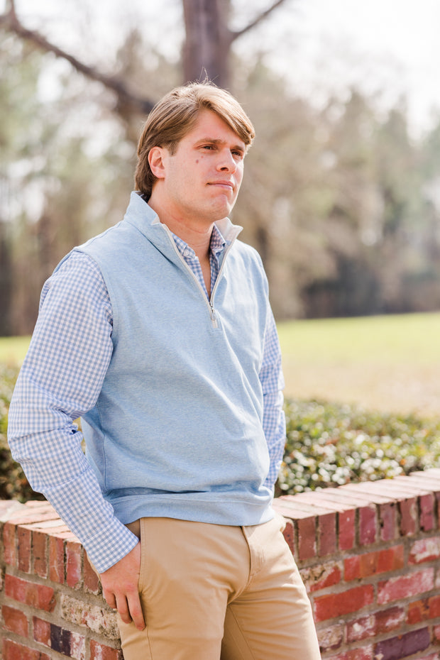 A man in a blue vest leaning against a brick wall wearing a Henning Shirt: St. Charles woven shirt.