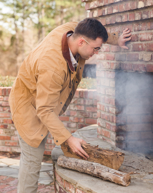 A man wearing a Bluff Barn Jacket is putting wood in a brick fireplace.