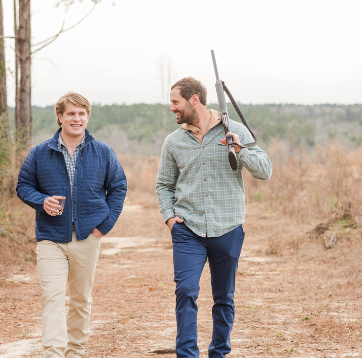 Two men wearing tailored fit flat-front Thomasville pants are walking through the woods in Thomasville, carrying rifles.