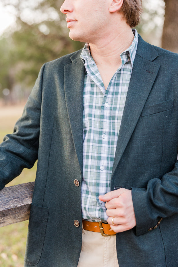 A Classic Unlined Gravier Gentleman's Jacket in a green blazer, leaning against a fence.